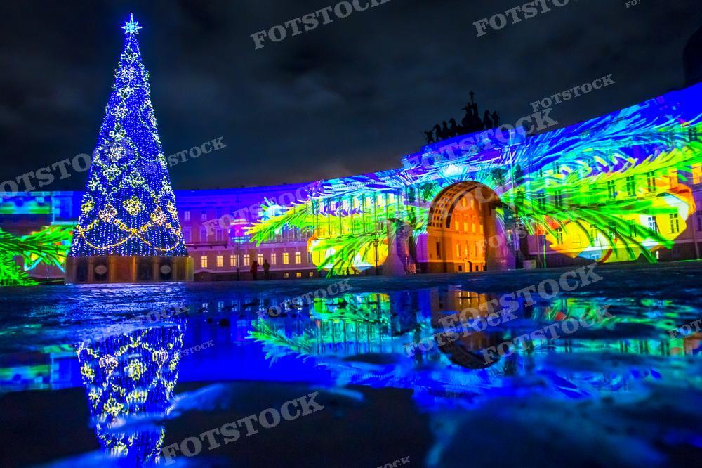 На фотографии представлен новогодний пейзаж: ярко украшенная Рождественская ёлка стоит на фоне здания, освещённого красочным световым шоу. Отражения украшений ёлки и цветных проекций на здании играют на мокрой поверхности, создавая ощущение сказочности и праздничной атмосферы. Это фото может использоваться для рекламы зимних праздников, поздравительных открыток, а также в качестве иллюстрации в статьях о новогодних мероприятиях в городе.